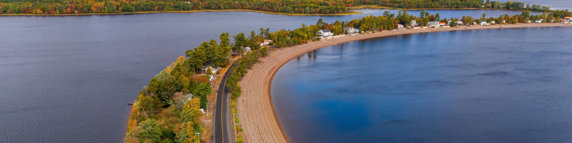 An aerial view of a curving road alongside a large body of water with a forested shoreline and a small settlement.
