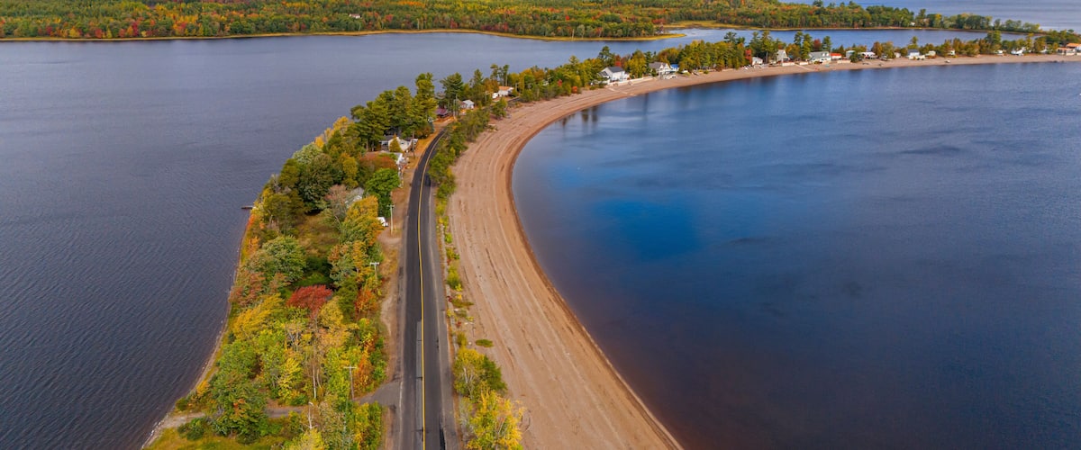 An aerial view of a curving road alongside a large body of water with a forested shoreline and a small settlement.