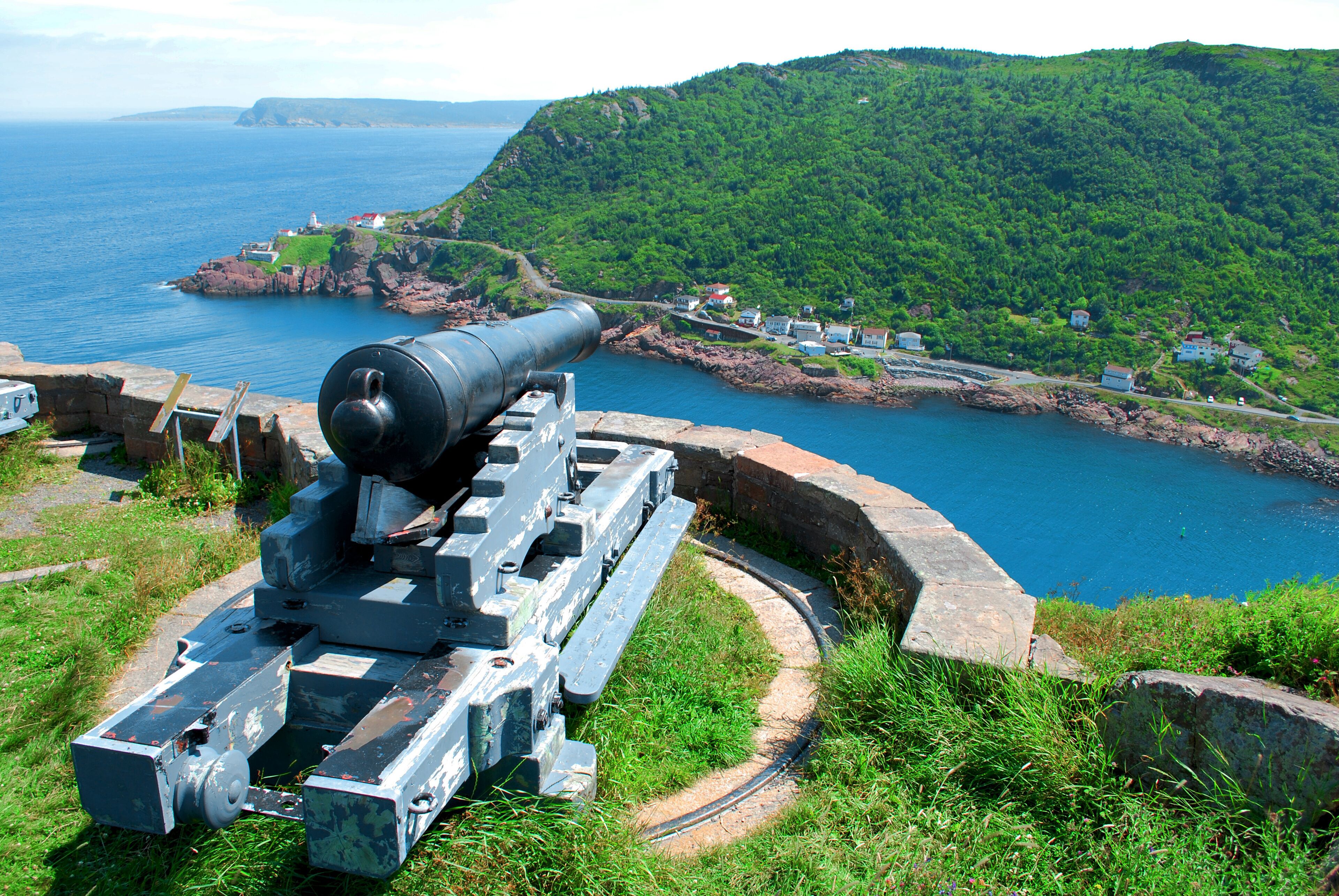 A coastal cannon on a grassy hill overlooking a bay with houses and cliffs.
