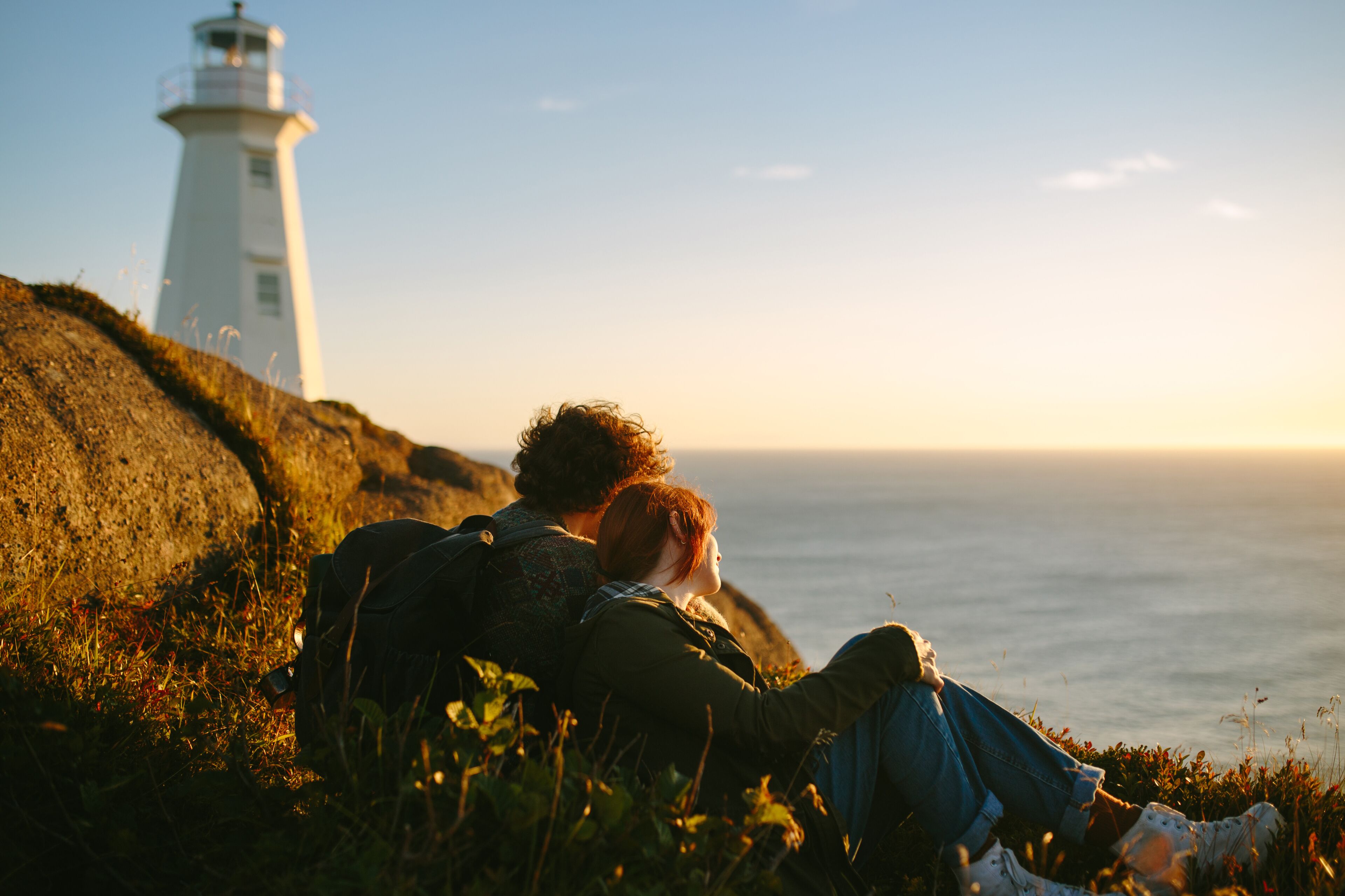 A couple sitting on a grassy cliff overlooking the ocean with a lighthouse in the background.