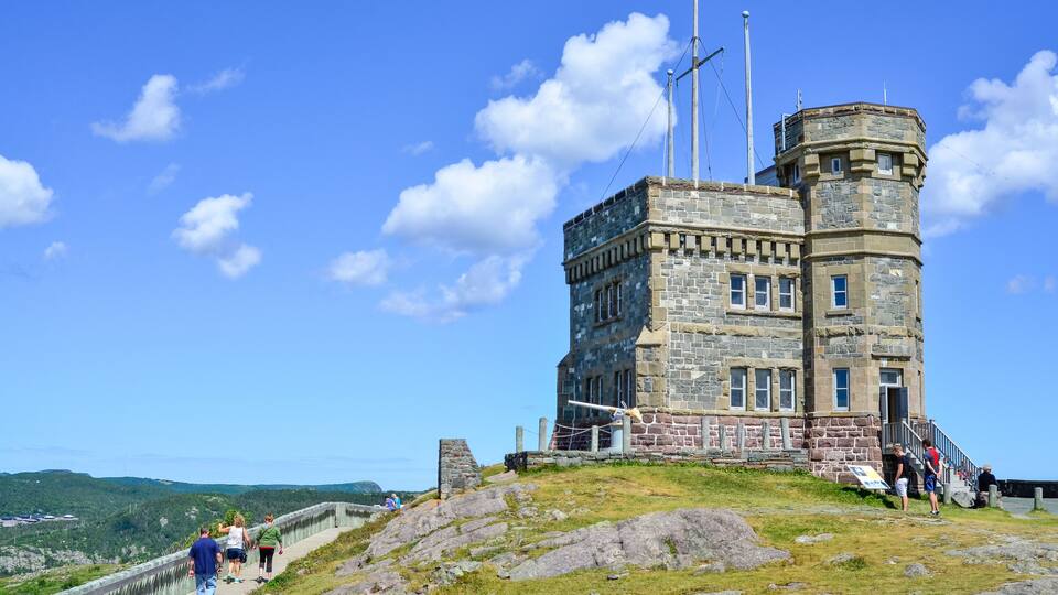 A stone tower with a flagpole, surrounded by a stone wall and grassy terrain.