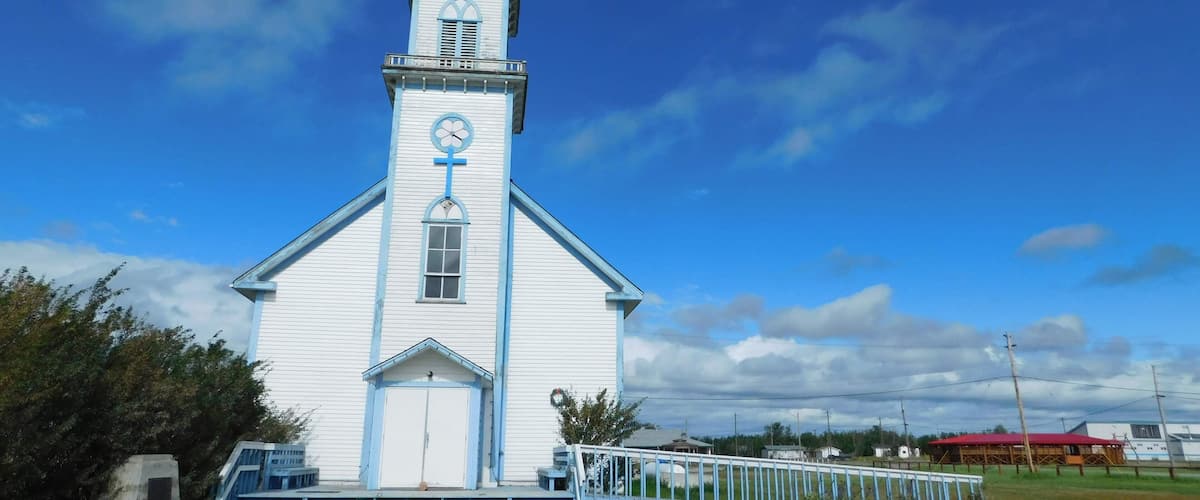 A white church with a blue cross and clock on the tower, surrounded by grass and a clear sky.