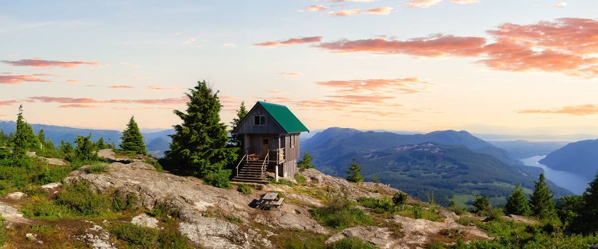 A wooden cabin with a green roof on a mountain ridge, surrounded by evergreen trees and a view of a valley with a lake.