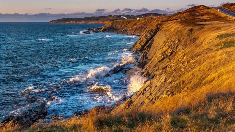 A coastal landscape with waves crashing against rocky cliffs and a road leading along the coast.