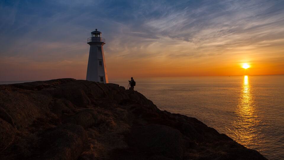 A lighthouse on a rocky coastline with the sun setting over the ocean.