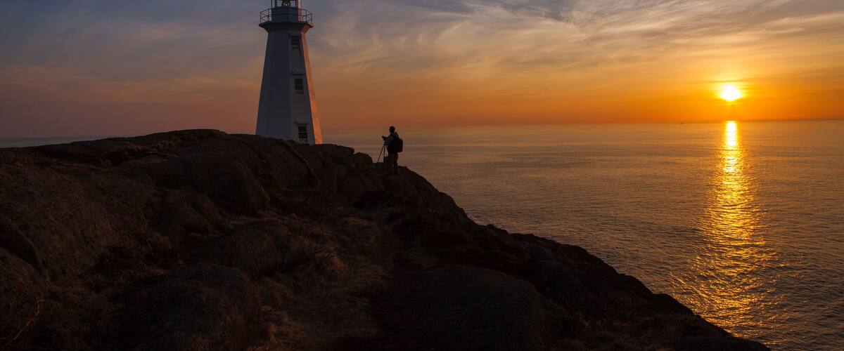 A lighthouse on a rocky coastline with the sun setting over the ocean.