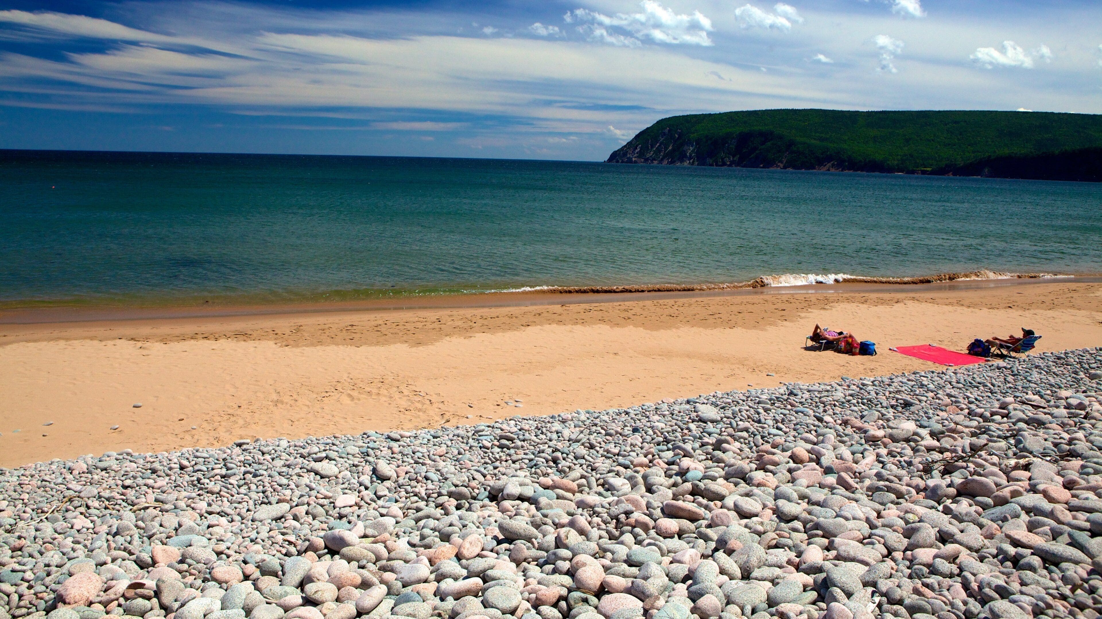 A pebbly beach with a red mat, a person lying on the sand, and a chair.