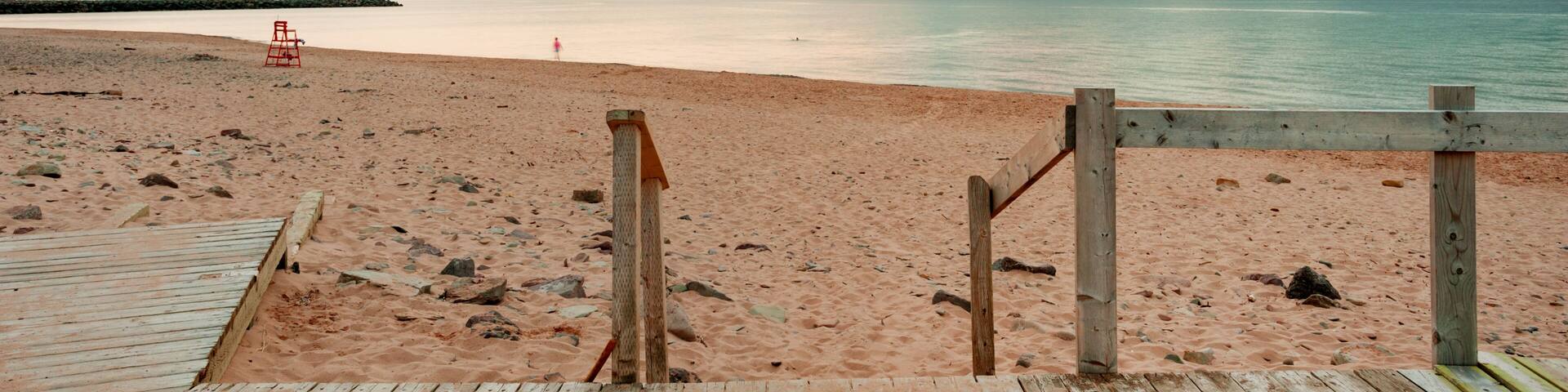 A wooden boardwalk leading to a sandy beach with a lifeguard chair and a rocky shoreline.