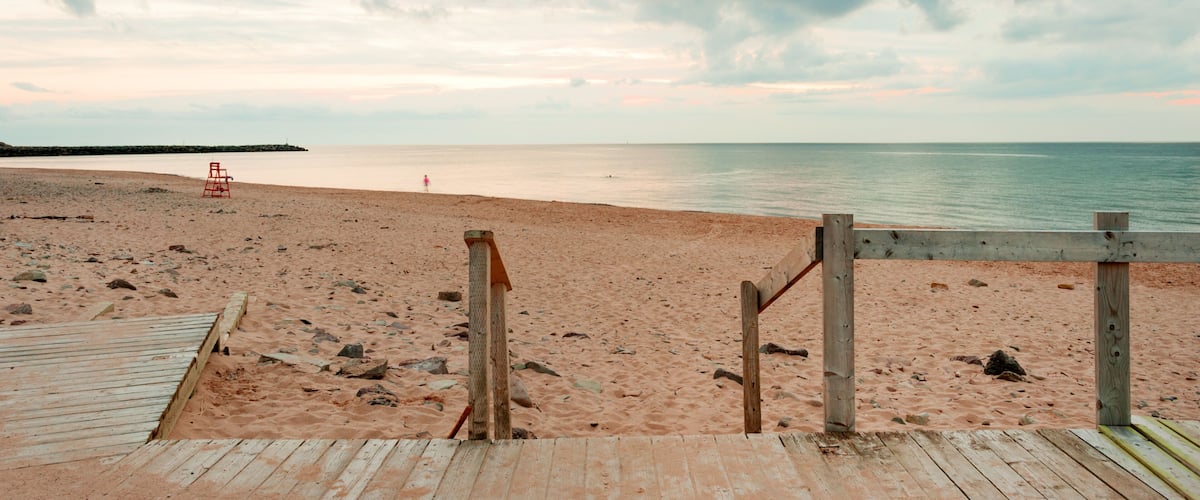A wooden boardwalk leading to a sandy beach with a lifeguard chair and a rocky shoreline.
