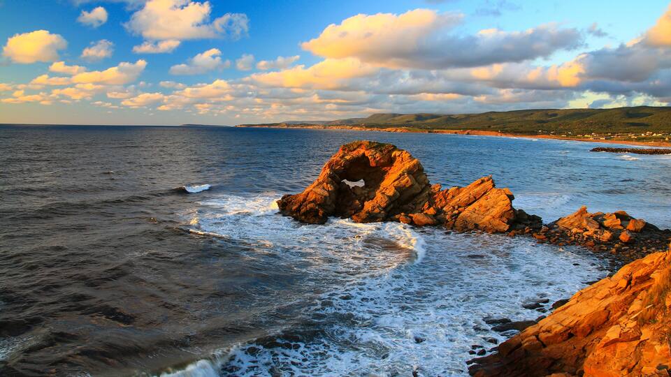 A rocky coastline with waves crashing against the shore and a clear sky with clouds.