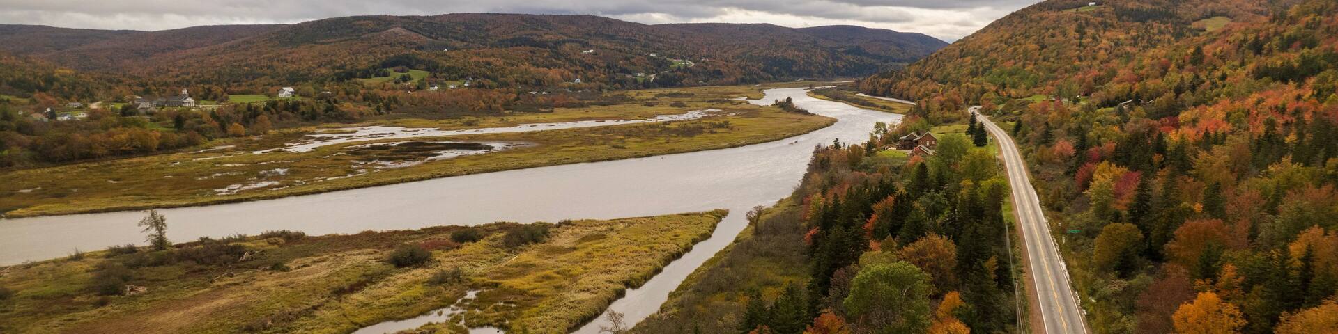 A river winding through a forested valley with autumn foliage, a road, and distant hills.