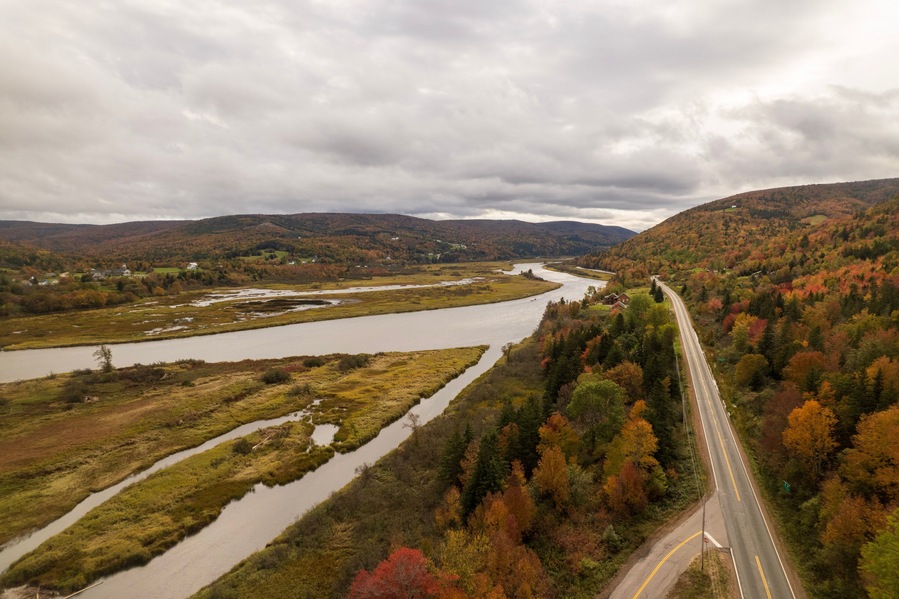 A river winding through a forested valley with autumn foliage, a road, and distant hills.