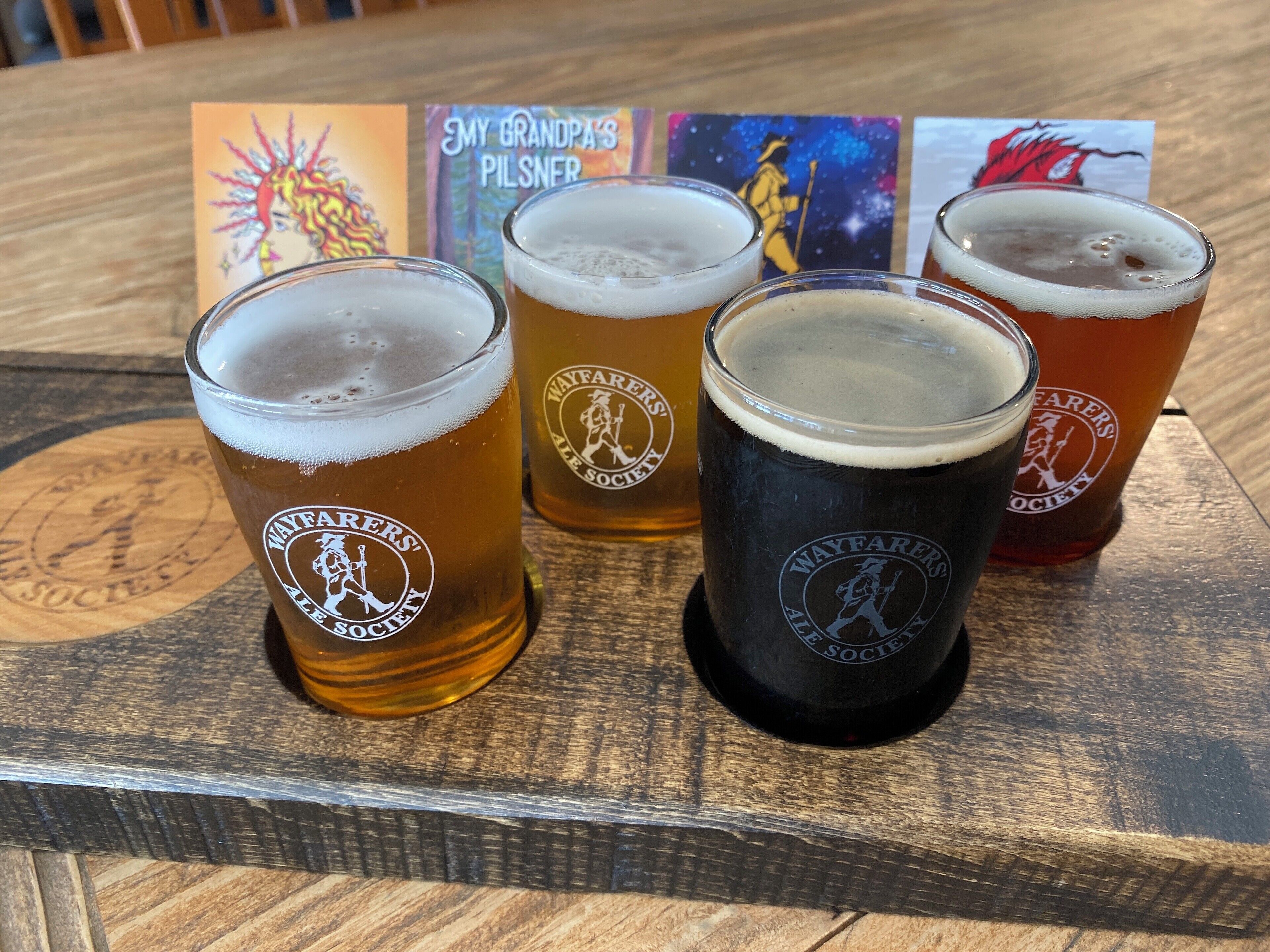 Four glasses of beer on a wooden tray with Wayfarers' Ale Society branding.