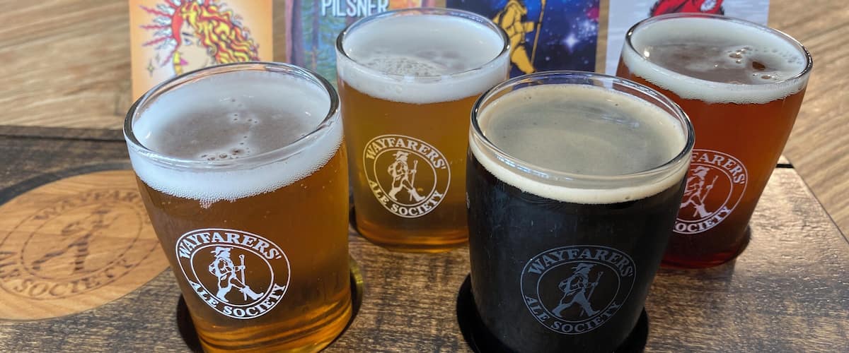 Four glasses of beer on a wooden tray with Wayfarers' Ale Society branding.