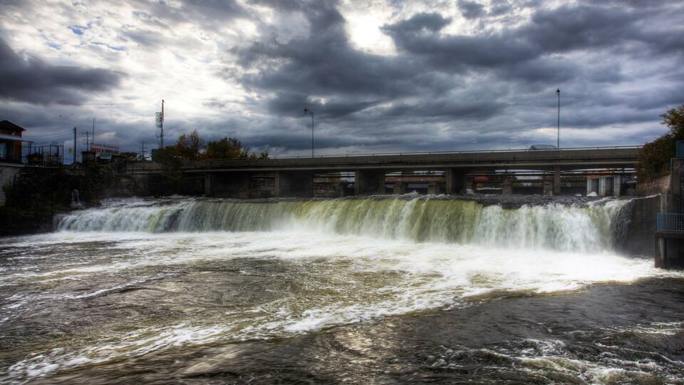 A waterfall cascading over a concrete barrier with a bridge in the background.