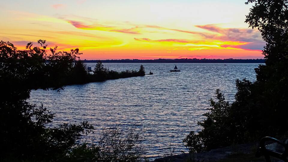 A sunset over a calm river with boats, surrounded by trees and a distant shoreline.