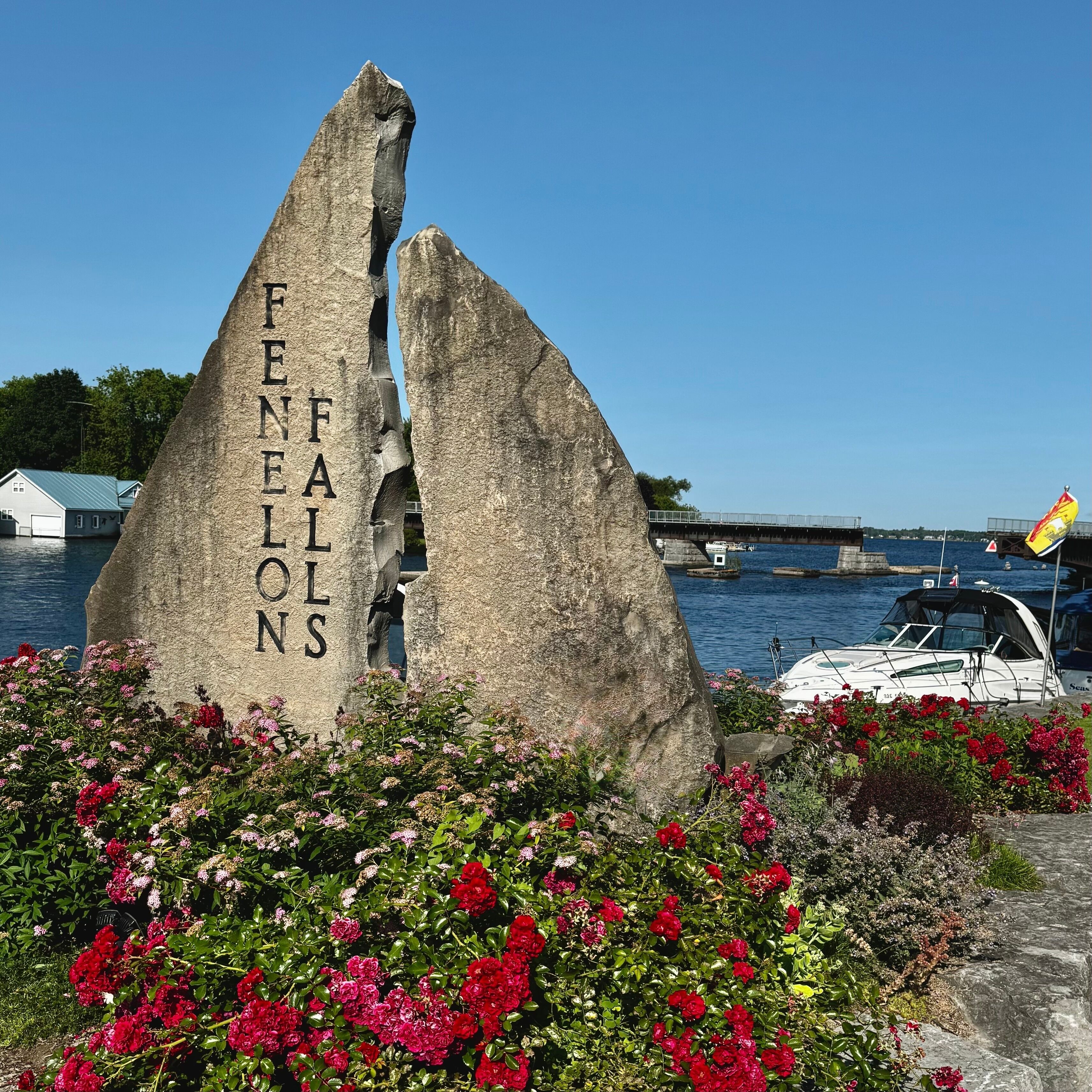 A rock with 'FENELONS' carved on it, surrounded by flowers and a body of water with boats.