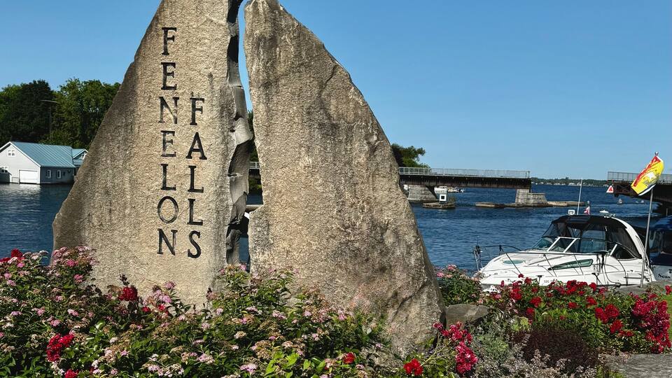 A rock with 'FENELONS' carved on it, surrounded by flowers and a body of water with boats.