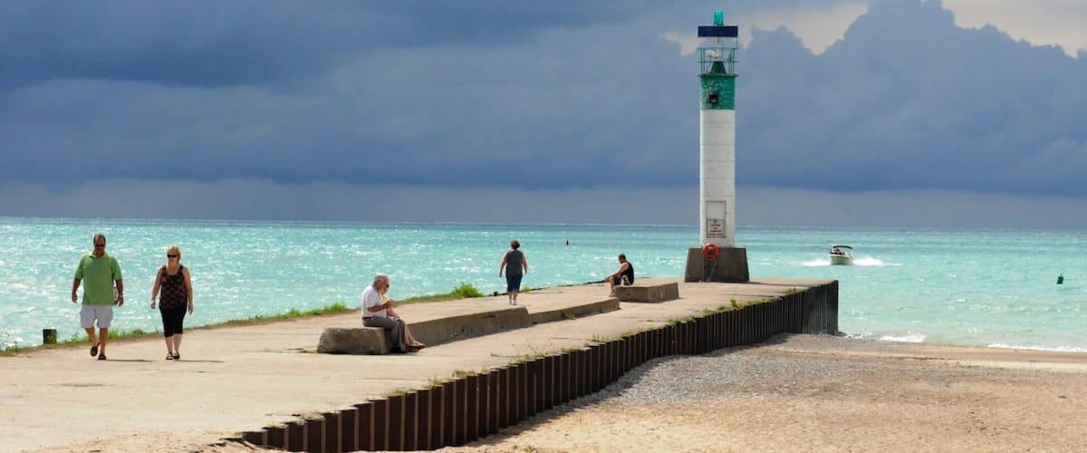 A coastal scene with a lighthouse, people walking, and a boat on the water.