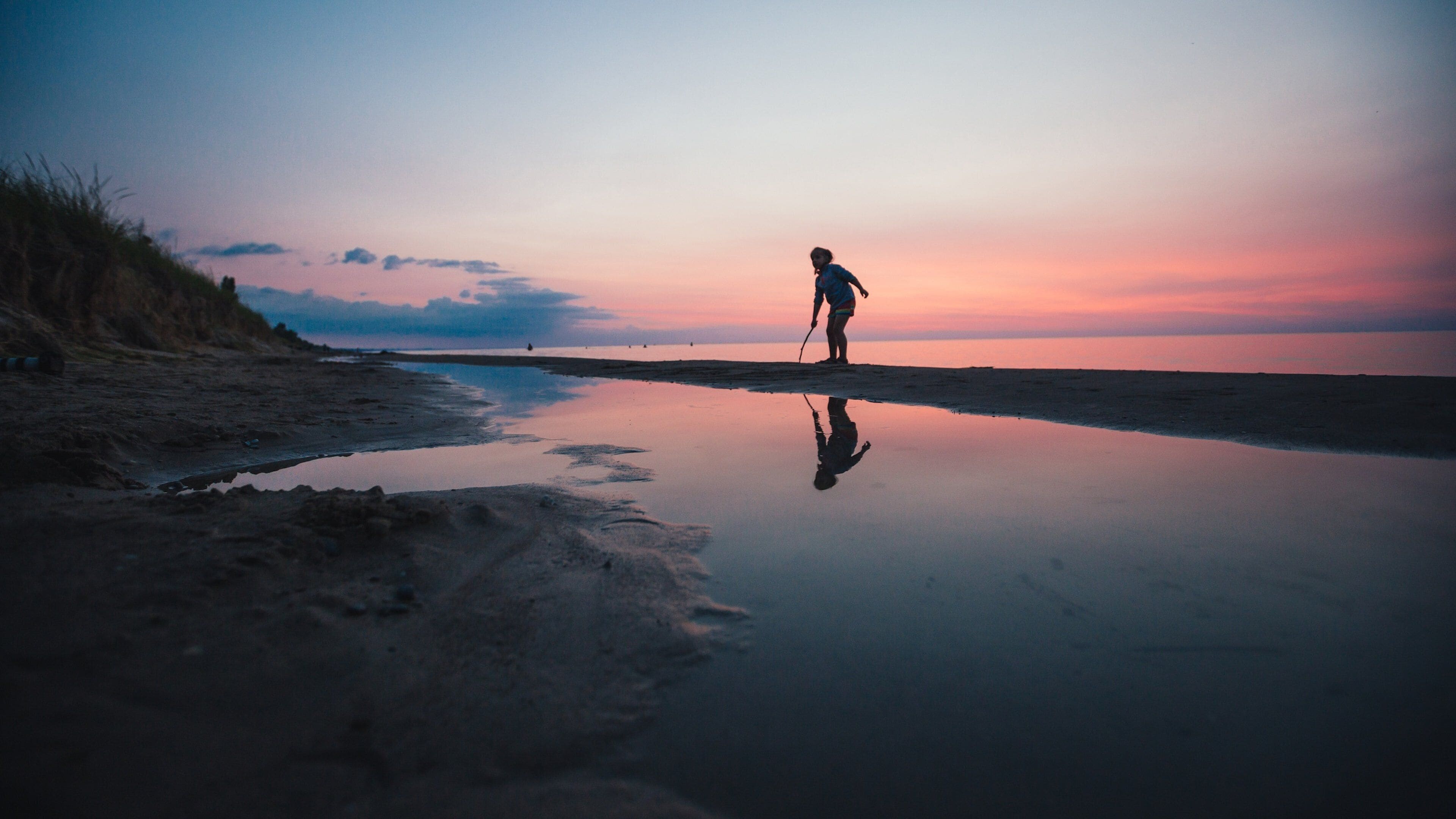 Una persona caminando en la playa al atardecer, con un mar tranquilo y un cielo despejado.