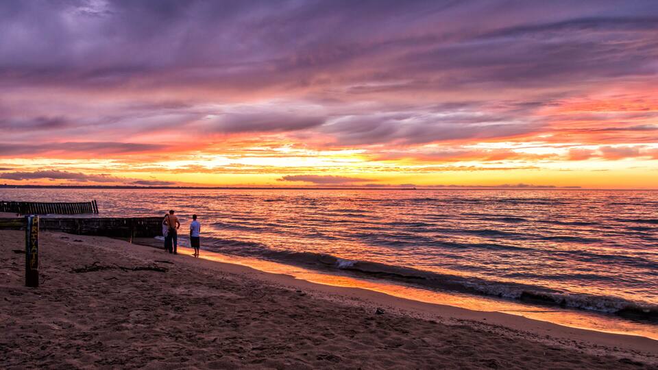 A beach with a sunset, two people standing by the water, and a wooden pier.