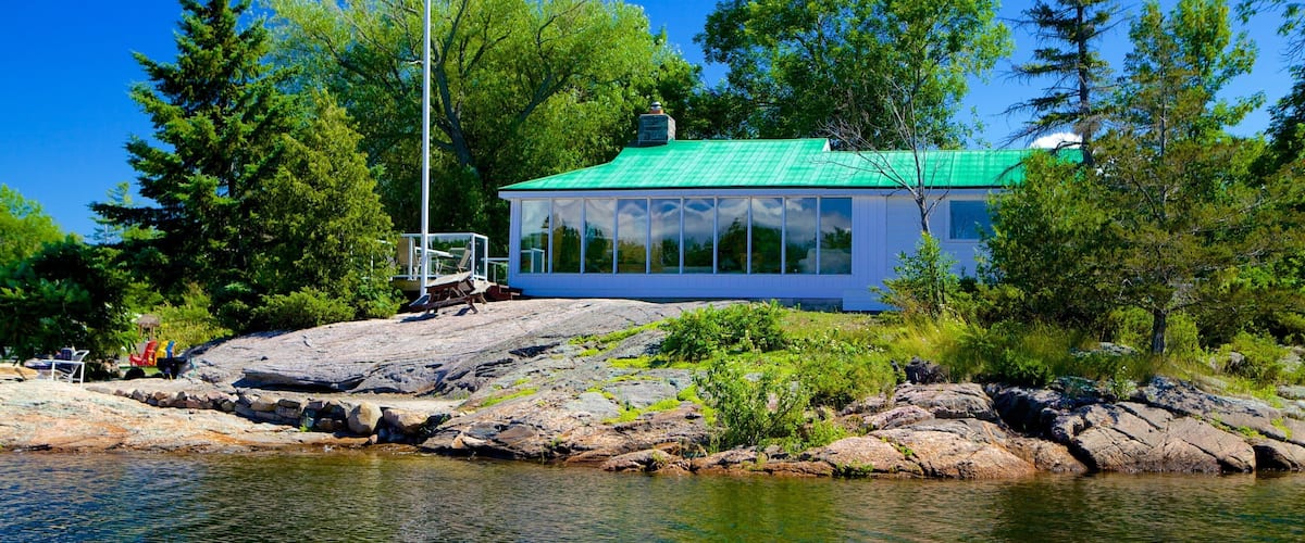 A cabin with a green roof on a rocky shore surrounded by trees.