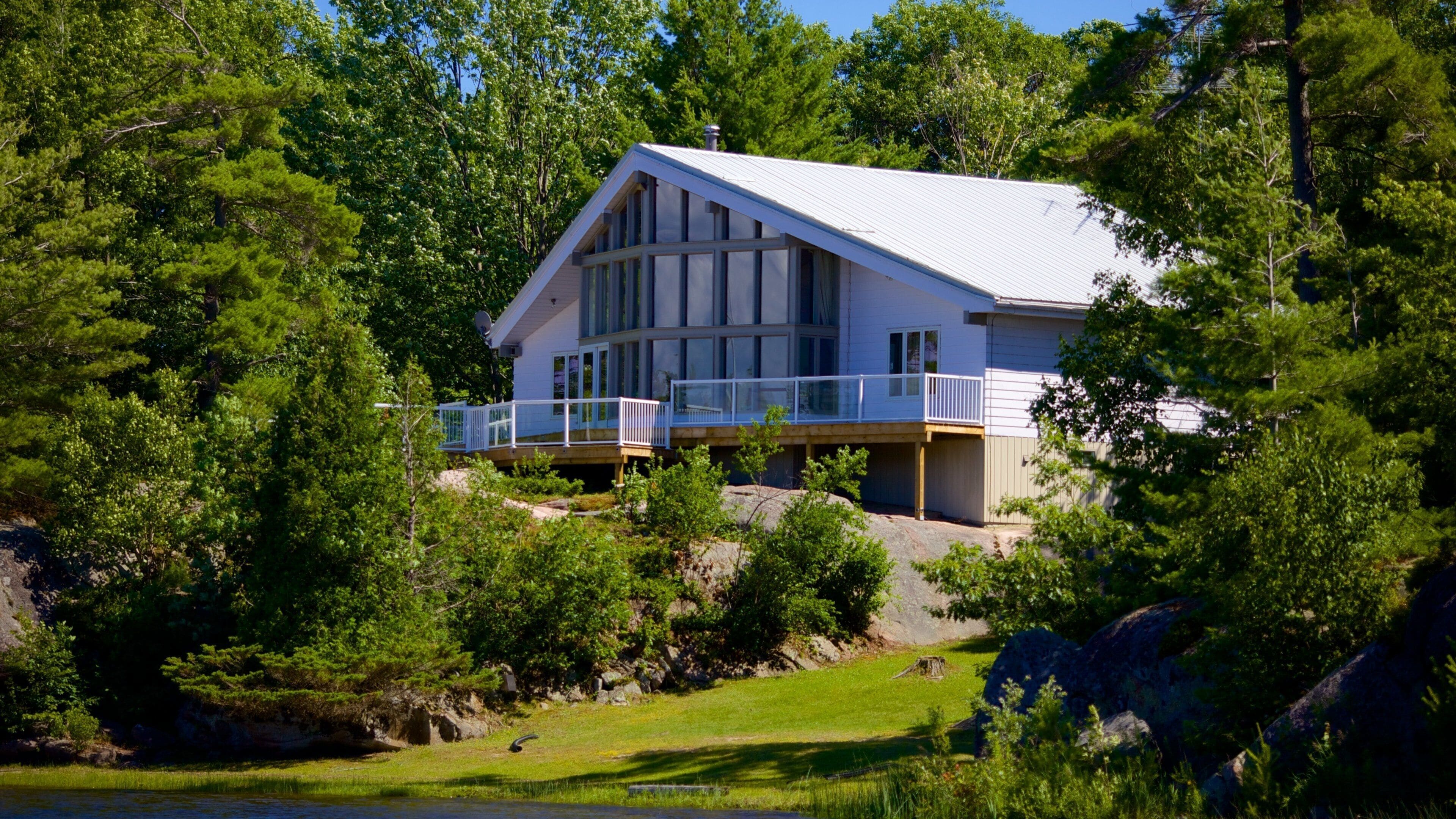 A large house with a white roof and a balcony, surrounded by lush green trees and a rocky landscape.