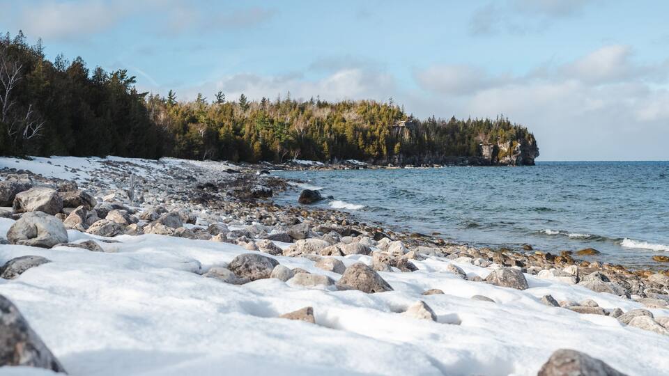 A rocky shoreline with snow-covered ground and a forested cliff in the background.