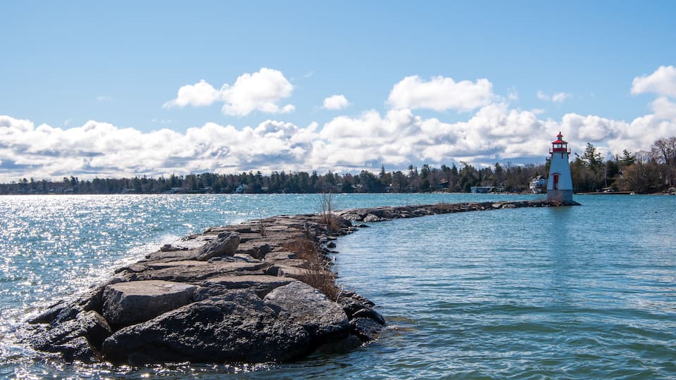A lighthouse on a rocky breakwater extending into a calm body of water, with a forested shoreline and a partly cloudy sky in the background.