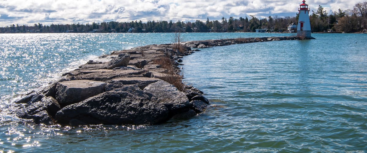 A lighthouse on a rocky breakwater extending into a calm body of water, with a forested shoreline and a partly cloudy sky in the background.