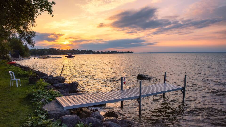 A wooden dock extending into calm water, surrounded by rocks and greenery, with a sunset in the background.