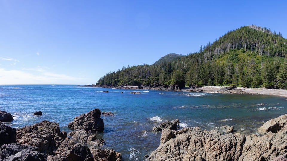 A rocky coastline with clear blue waters, a sandy beach, and a forested hill.