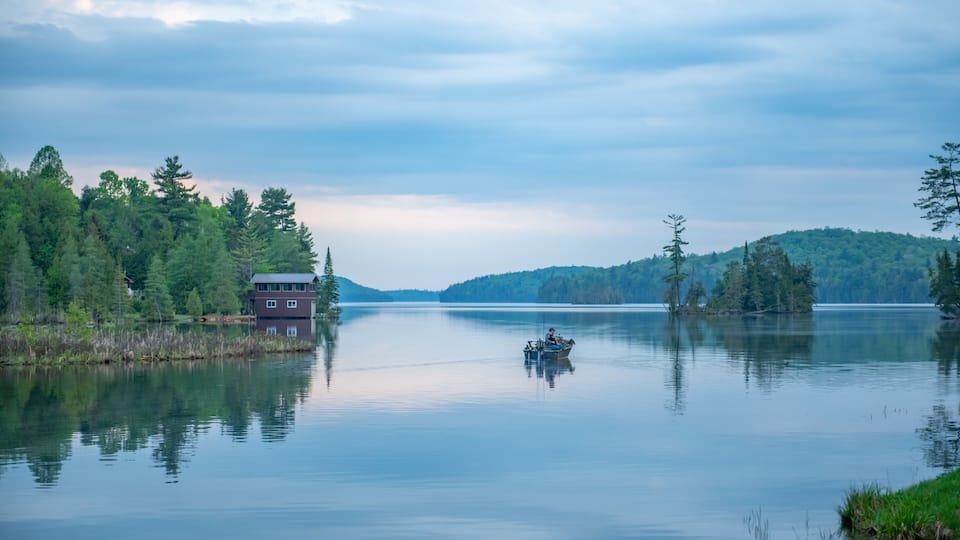 A calm lake with a small house on the shore, surrounded by dense trees and hills in the background.