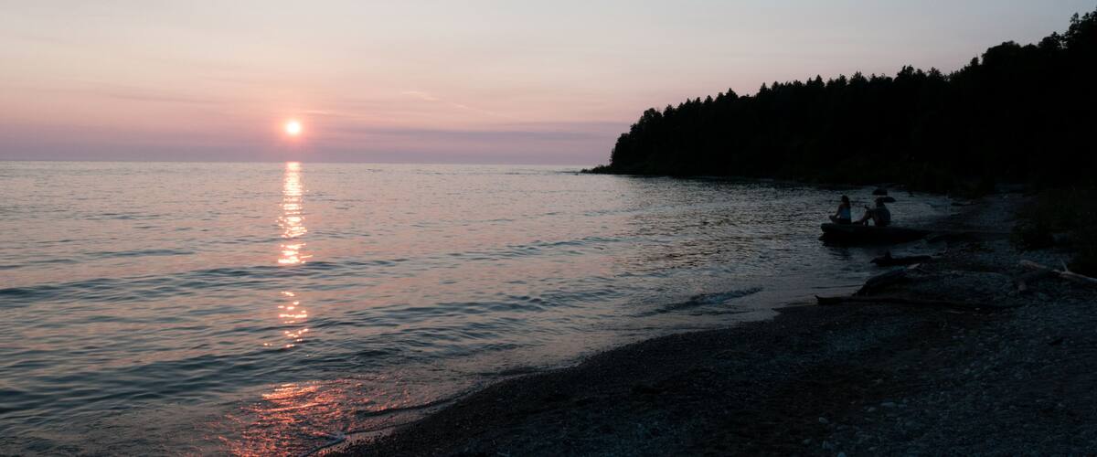 A sunset over a calm sea with a rocky shoreline and a forested hill in the background.