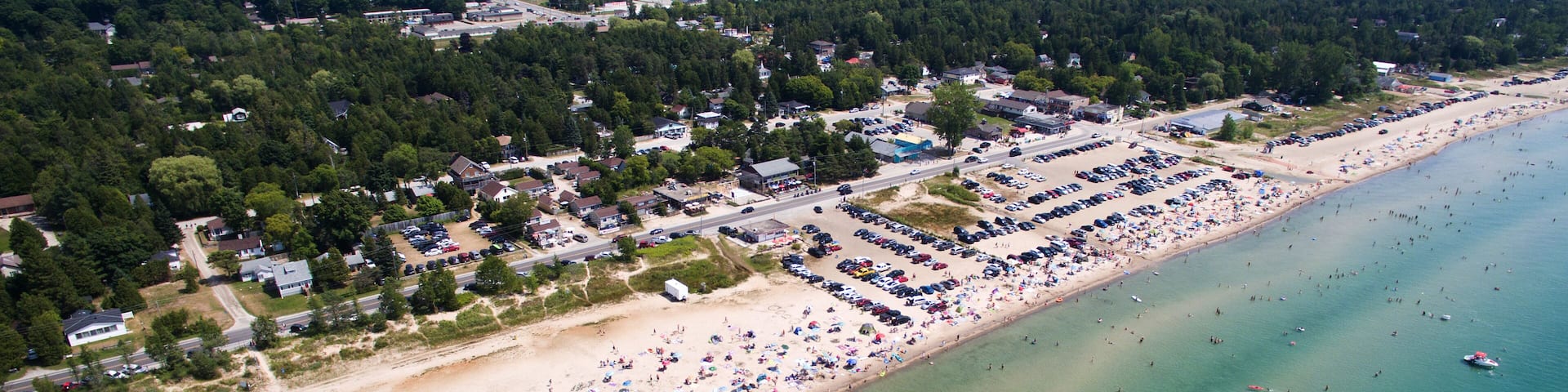A coastal town with a sandy beach, a parking lot, and a forested area in the background.