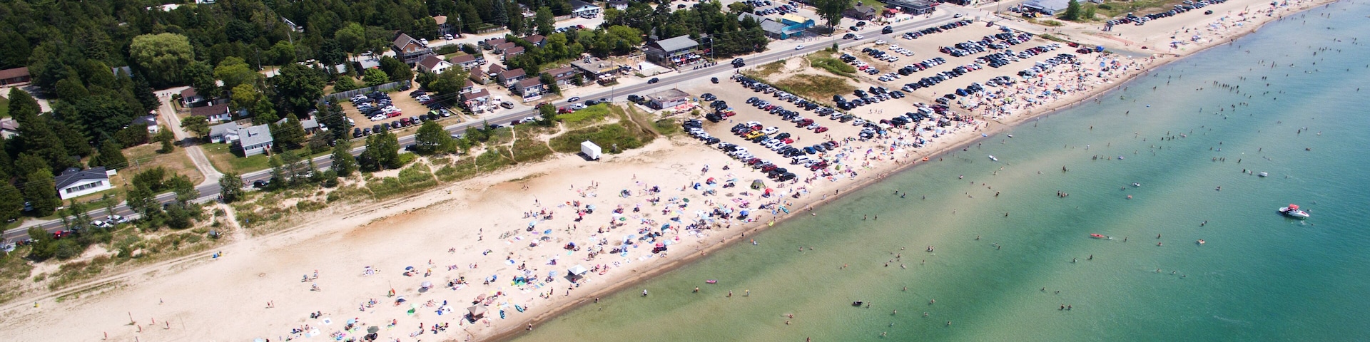 A coastal town with a sandy beach, a parking lot, and a forested area in the background.