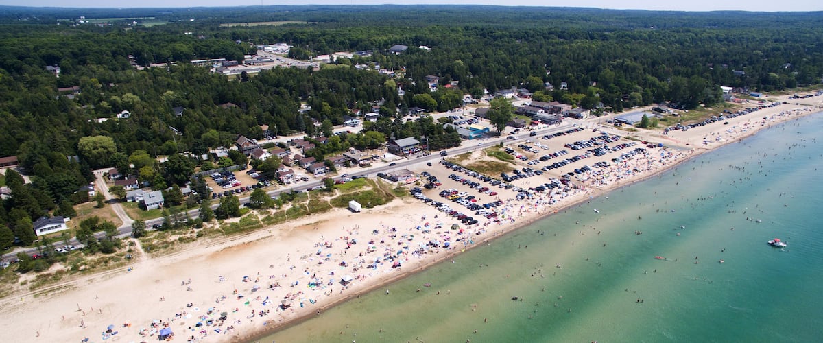 A coastal town with a sandy beach, a parking lot, and a forested area in the background.