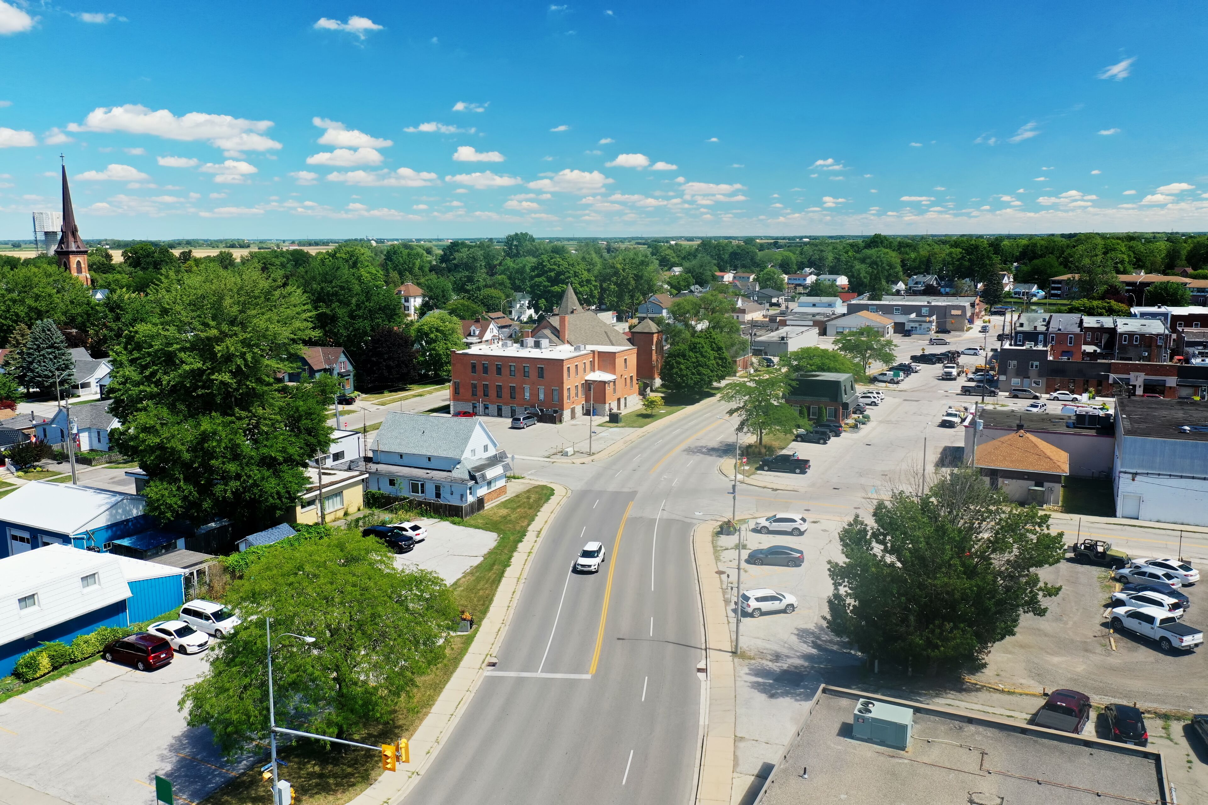 A town with a prominent church steeple, a large brick building, and a wide road with cars.