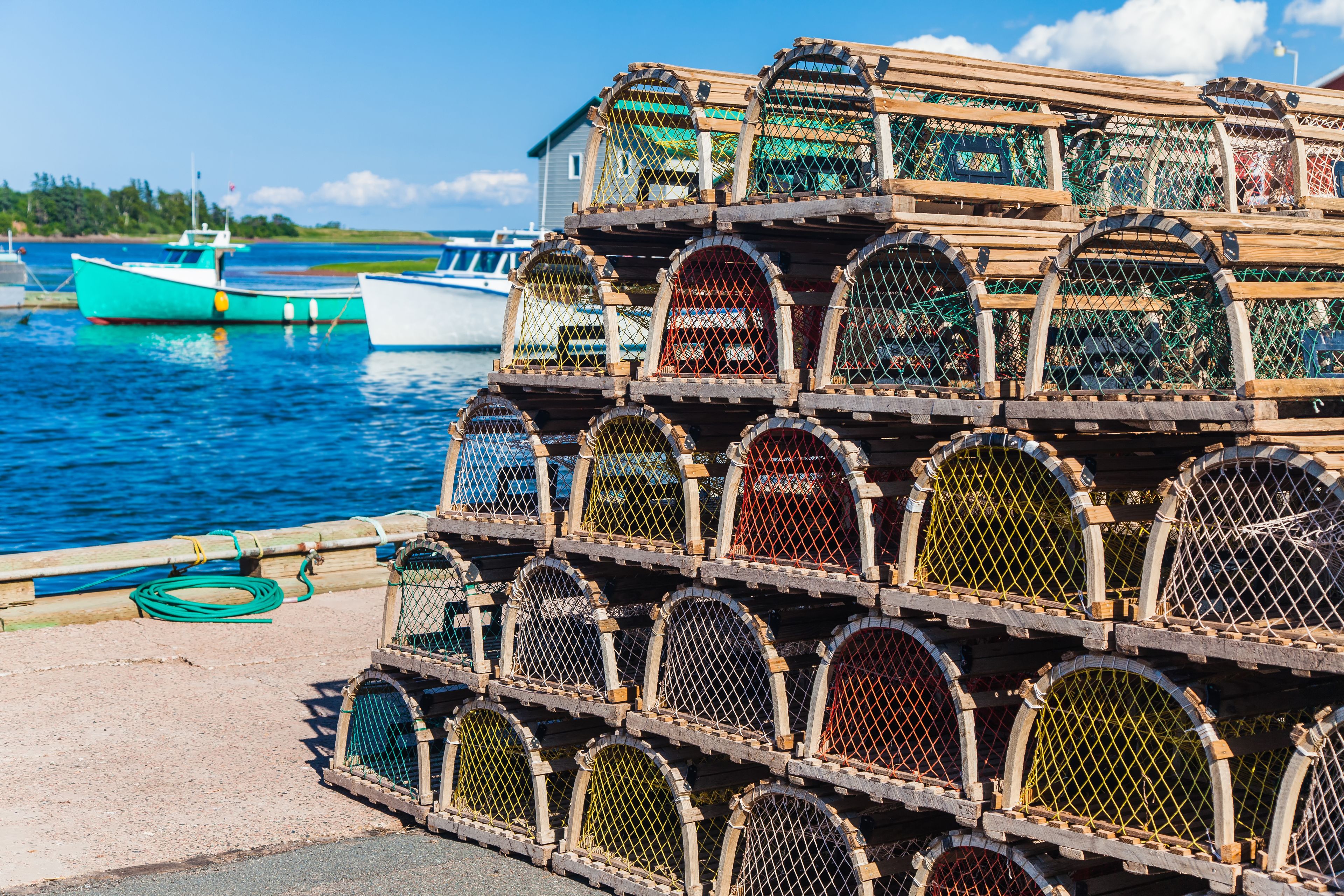 A stack of lobster traps by a waterfront with boats in the background.