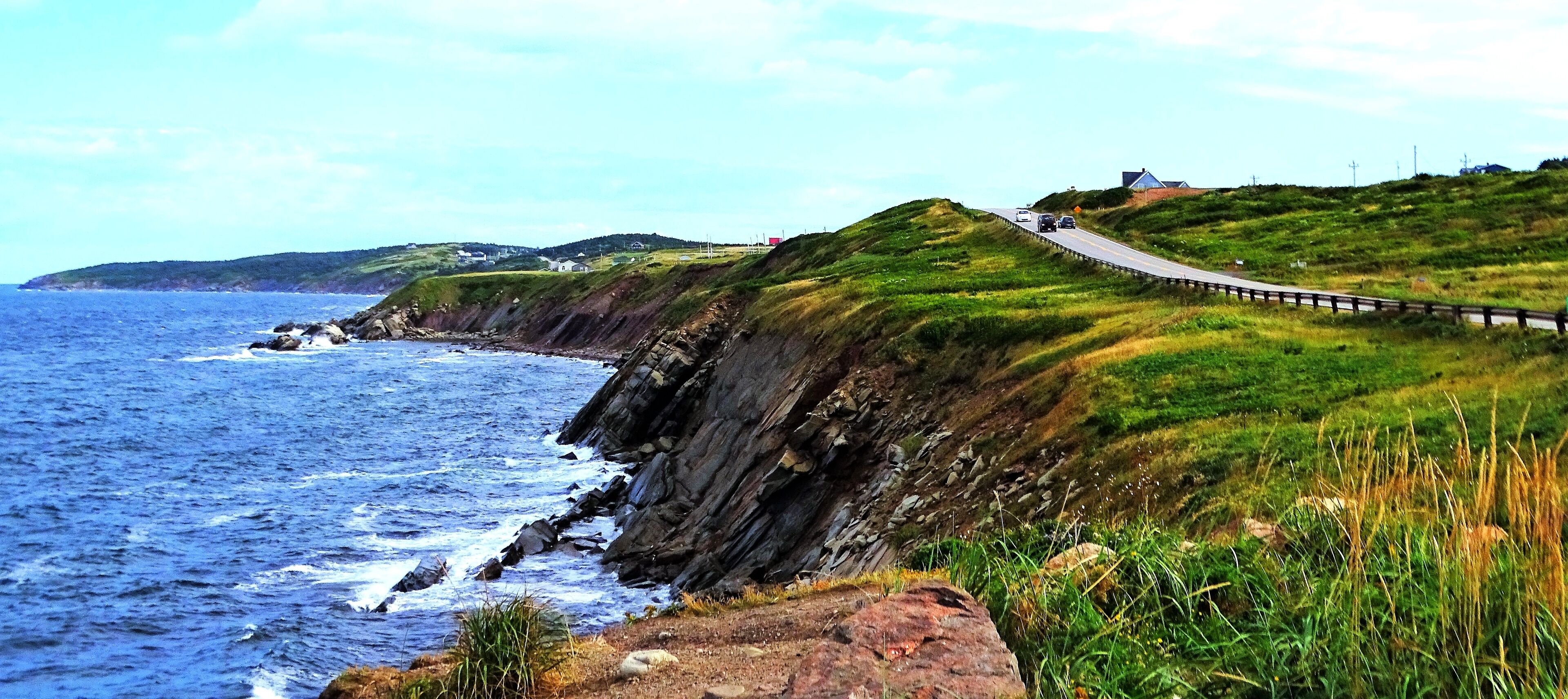 A coastal road with a guardrail, ocean waves, grassy hills, and distant houses.