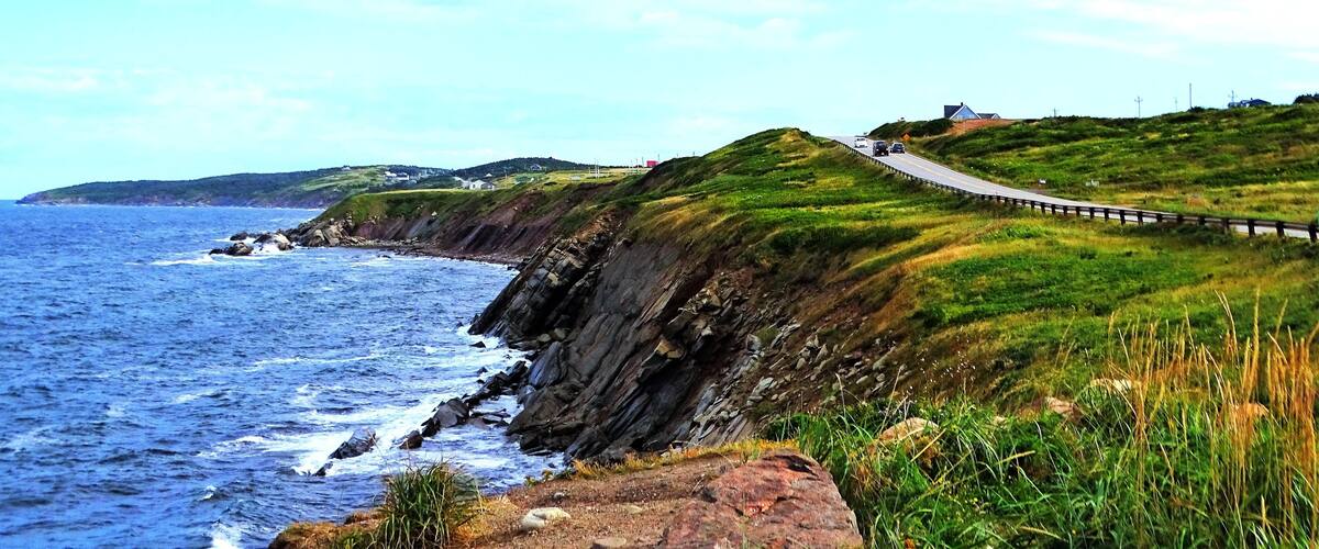 A coastal road with a guardrail, ocean waves, grassy hills, and distant houses.
