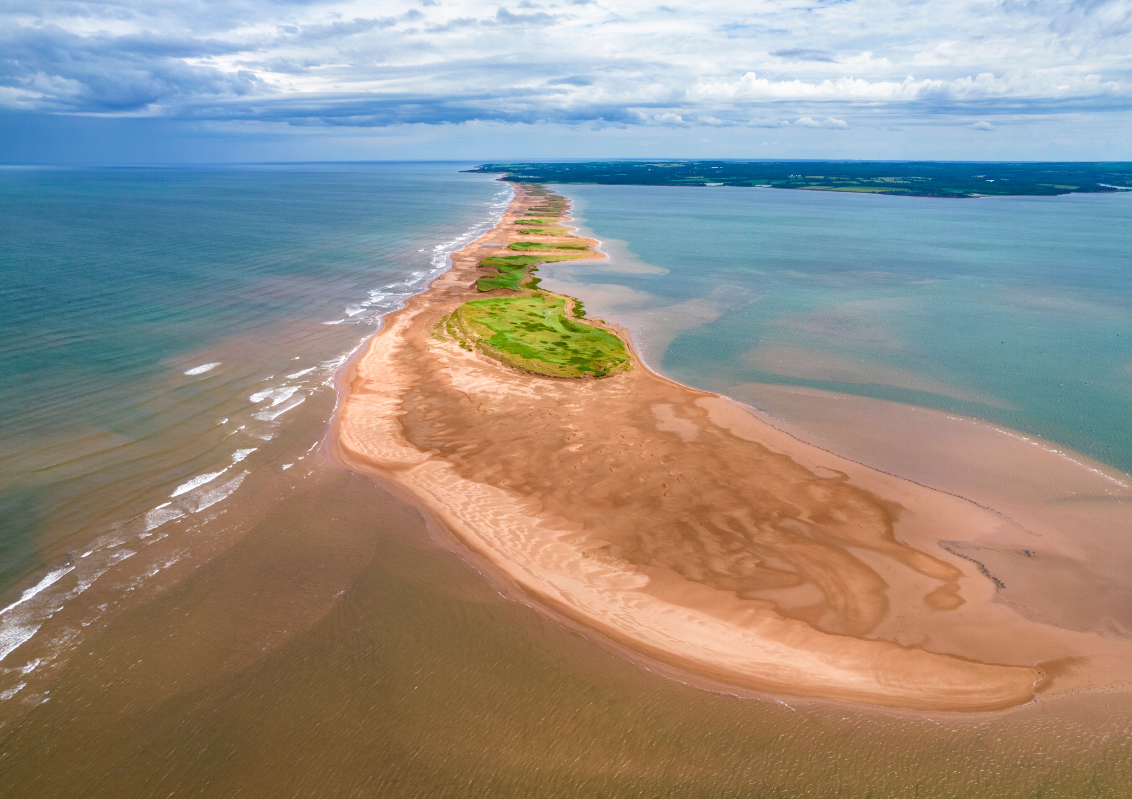 A coastal landscape with a long sandy beach, green vegetation patches, and a calm sea under a cloudy sky.