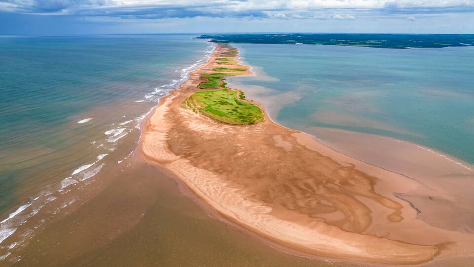 A coastal landscape with a long sandy beach, green vegetation patches, and a calm sea under a cloudy sky.