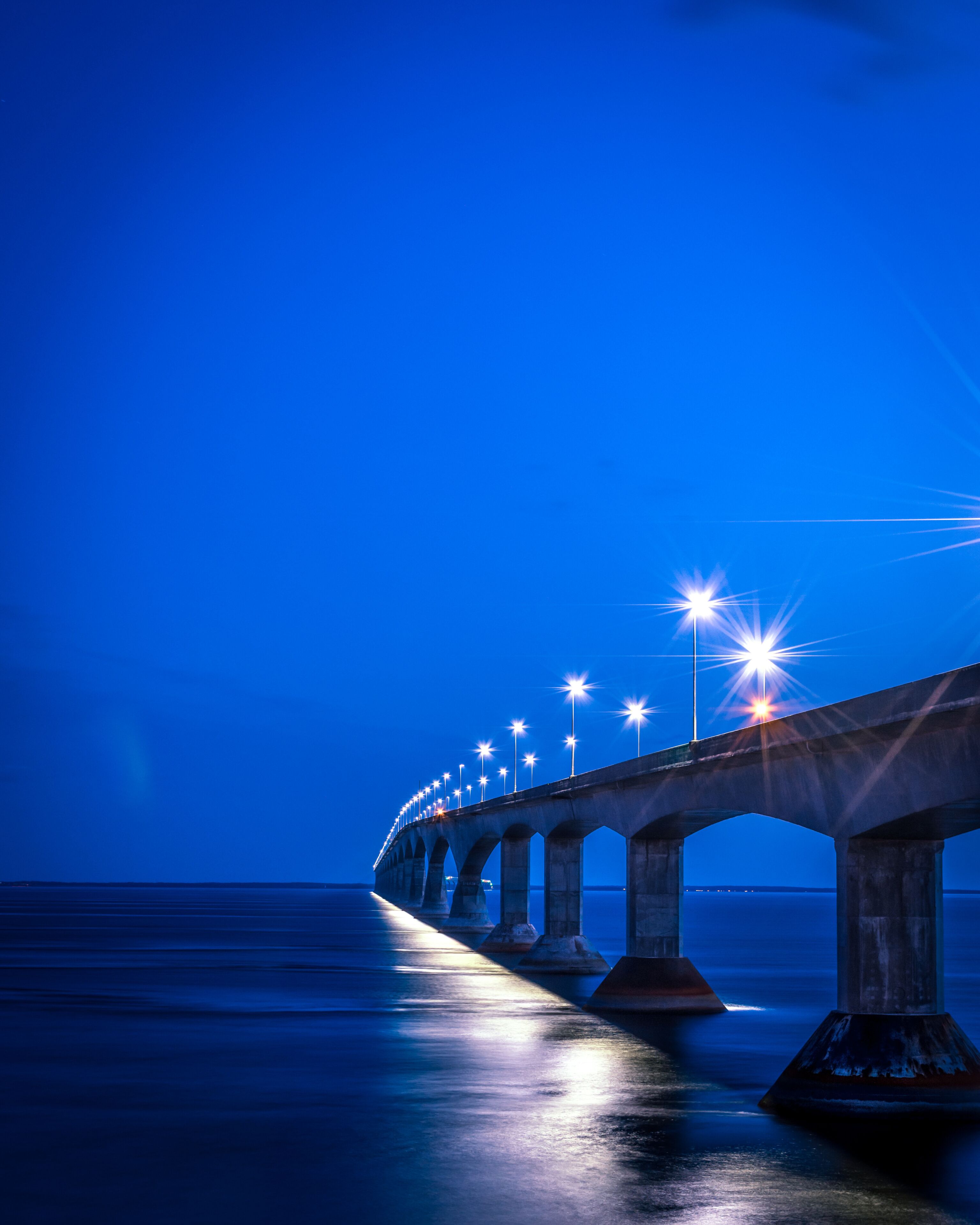 A long bridge with multiple arches, illuminated by streetlights, extending over a calm body of water at night.