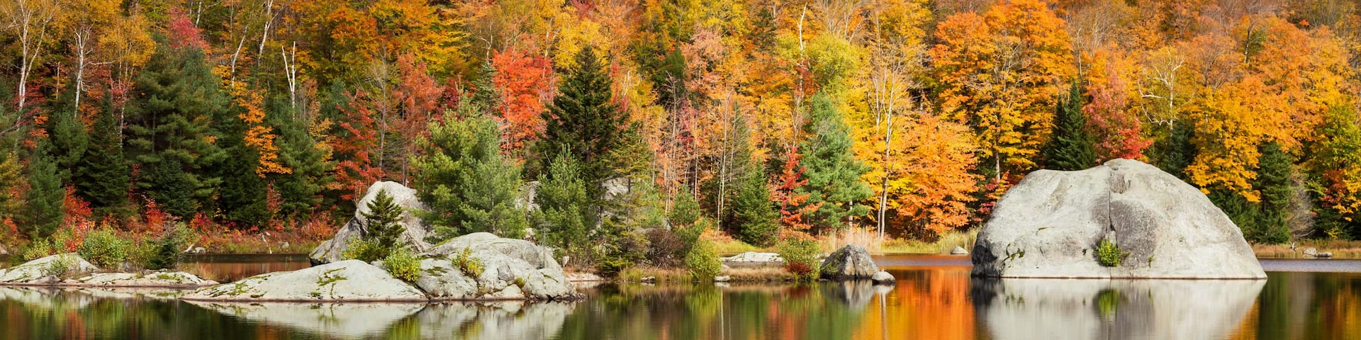 A calm lake reflecting autumn trees and a prominent rock formation.