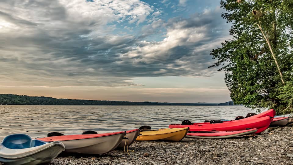 A row of kayaks on a pebbly shore with a lake and cloudy sky in the background.