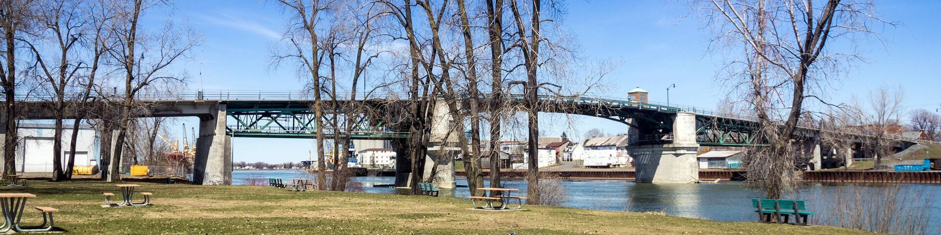 A park with benches and a river, a bridge, and leafless trees.