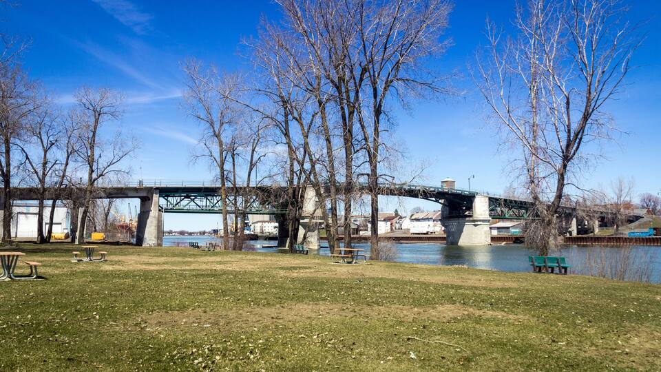 A park with benches and a river, a bridge, and leafless trees.