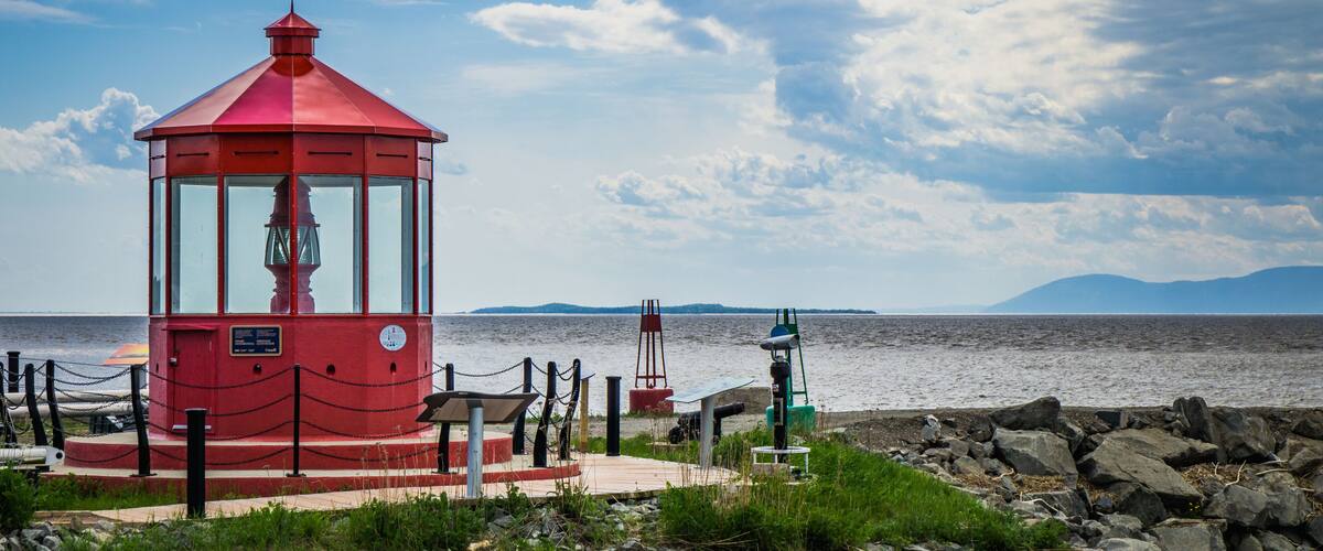 A red lighthouse by a rocky shore with a calm sea and distant mountains.