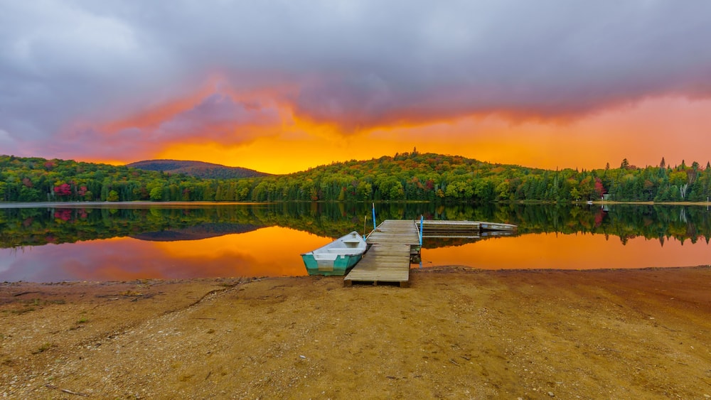A serene lake with a wooden dock and a small boat, surrounded by lush green trees and a dramatic sunset sky.